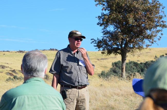 Ken Tate, wearing a baseball hat and sunglasses, addresses a group standing on grassy rangeland.