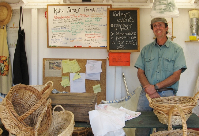 Farmer inside a farm stand surrounded by baskets and price board.